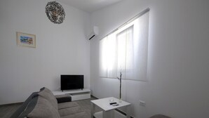 Un salon moderne avec un canapé gris, un téléviseur à écran plat posé sur un meuble blanc, une petite table blanche ornée d’un vase et une fenêtre avec des rideaux légers.