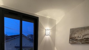 Un dormitori amb un llit, una tauleta de nit, una làmpada penjada a la paret, una finestra i un quadre.