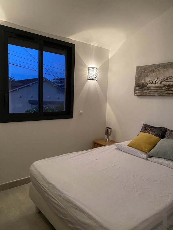 A bedroom with a bed, a nightstand, a wall-mounted lamp, a window, and a painting.