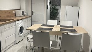 Cuina compacta amb rentadora, microones i nevera. Hi ha una taula de dinar amb cadires i una planta en test amb una maceta a la cantonada.