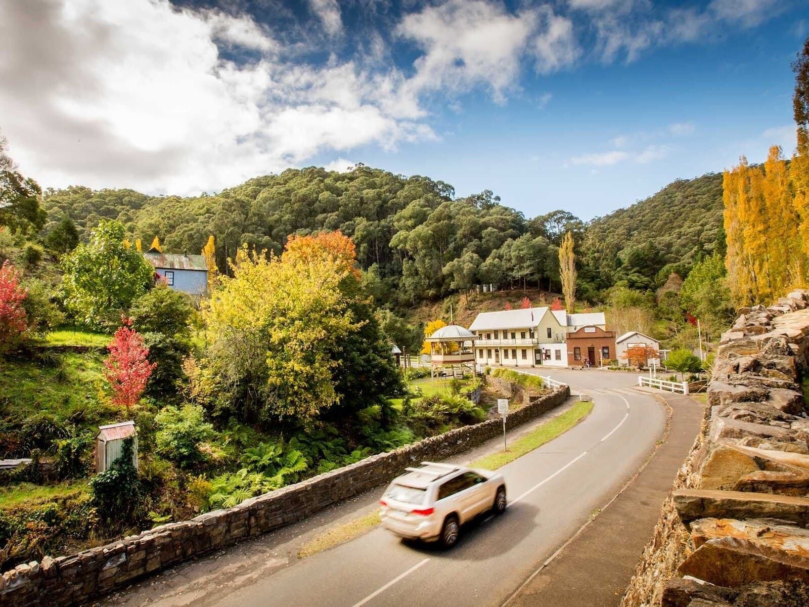 A white car driving on a road with a stone wall on one side and a building with a white roof and a signboard on the other.