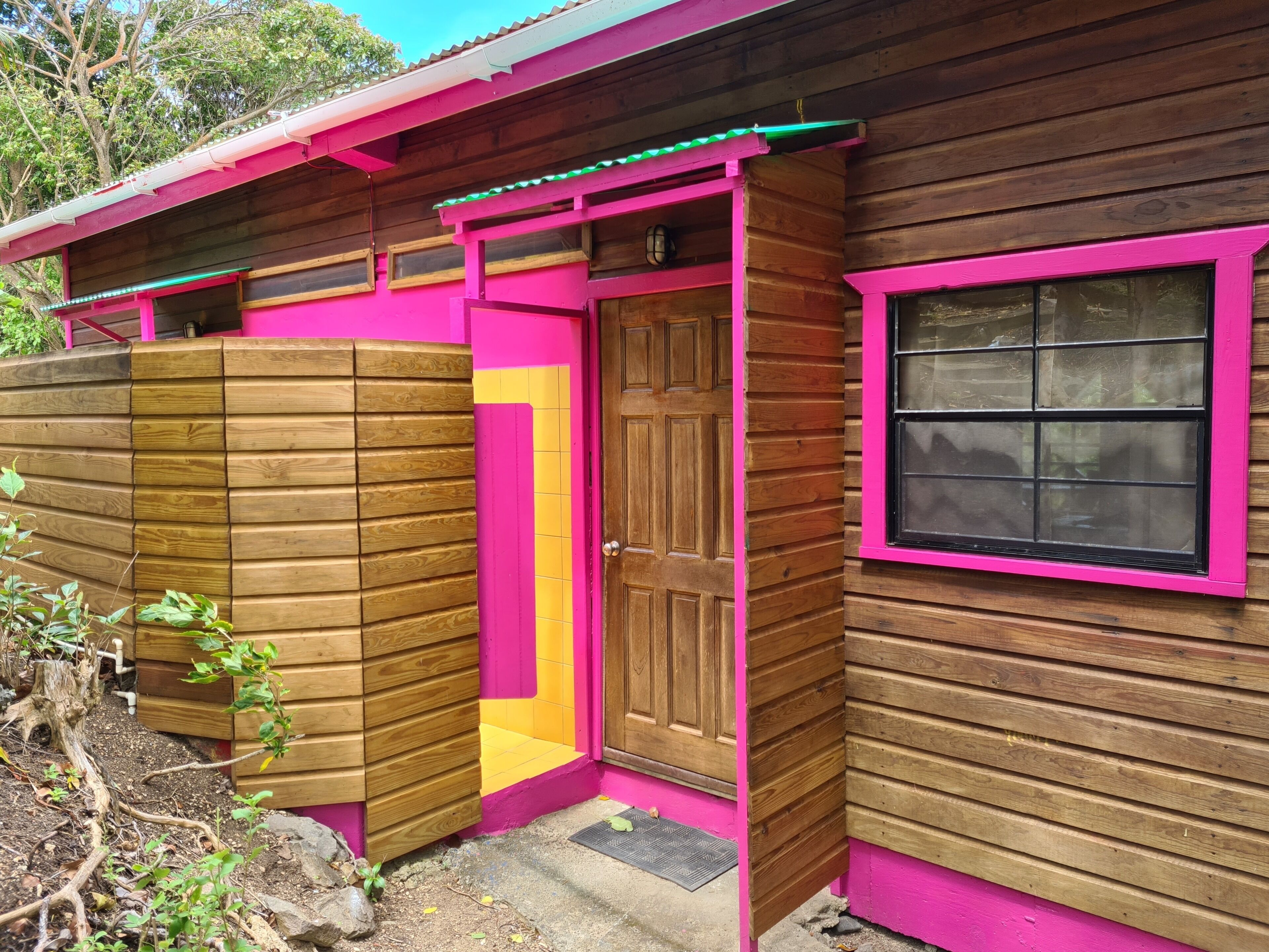 A wooden cabin with a pink door and window frames, a small porch area, and a wooden fence.