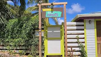 A wooden sign with "Welcome to Paradise" written on it, a white building with a yellow door, and lush green foliage in the background.