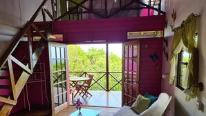 A cozy room with a wooden staircase, a glass table with a vase, a sofa, and a balcony with a view of greenery.