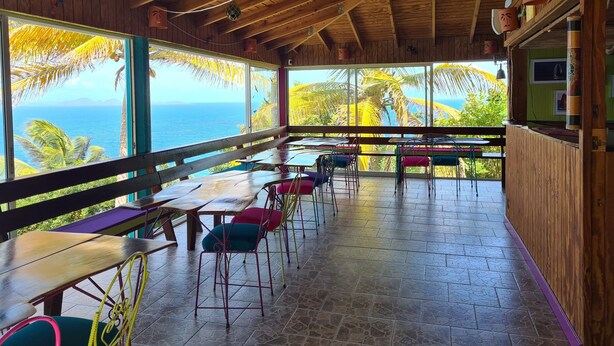 A restaurant with wooden beams and large windows overlooking the ocean. Tables and chairs are arranged for dining.