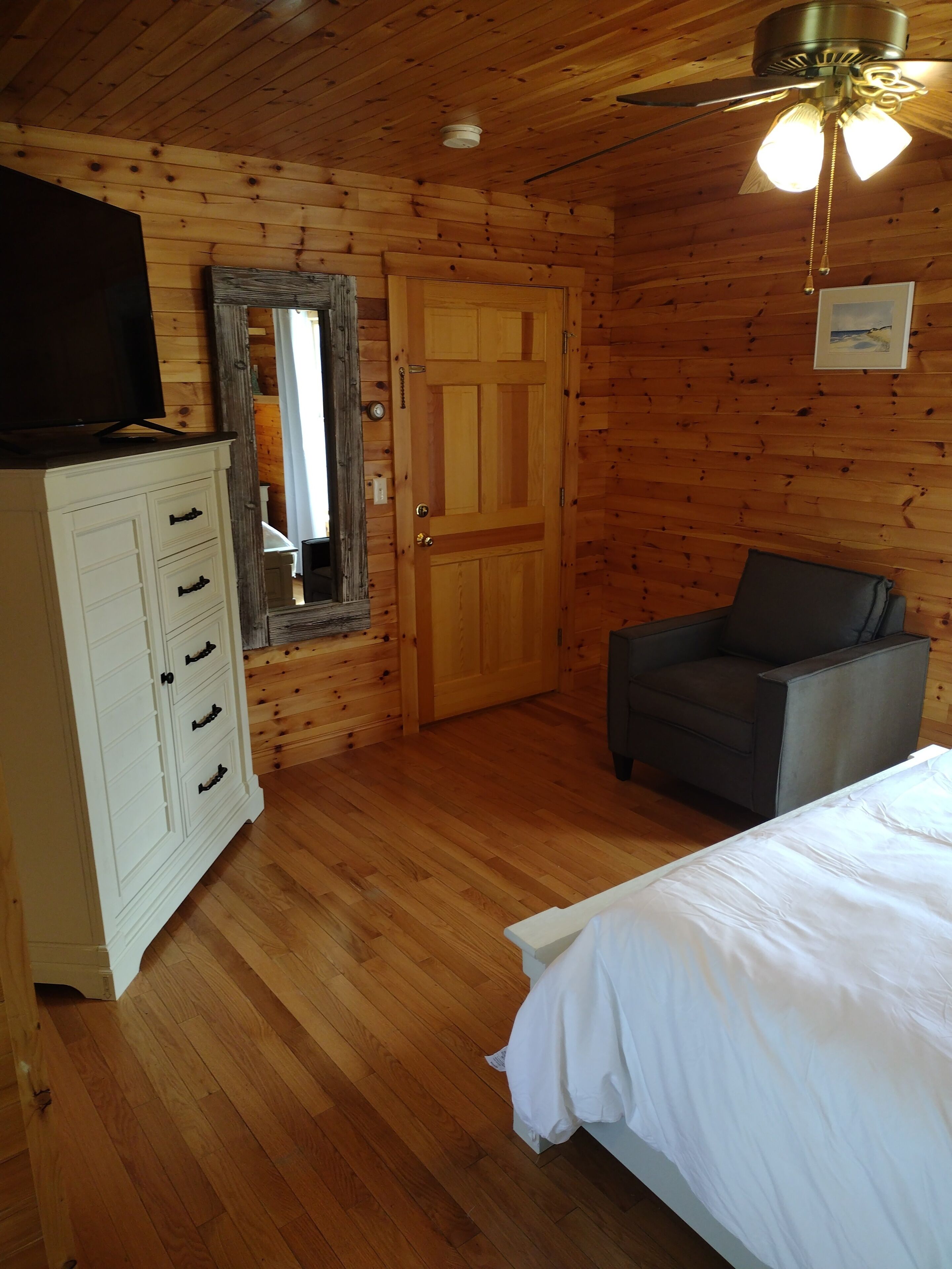 Una stanza con rivestimento in legno, un letto, una sedia, una cassettiera, uno specchio e una ventola a soffitto.
