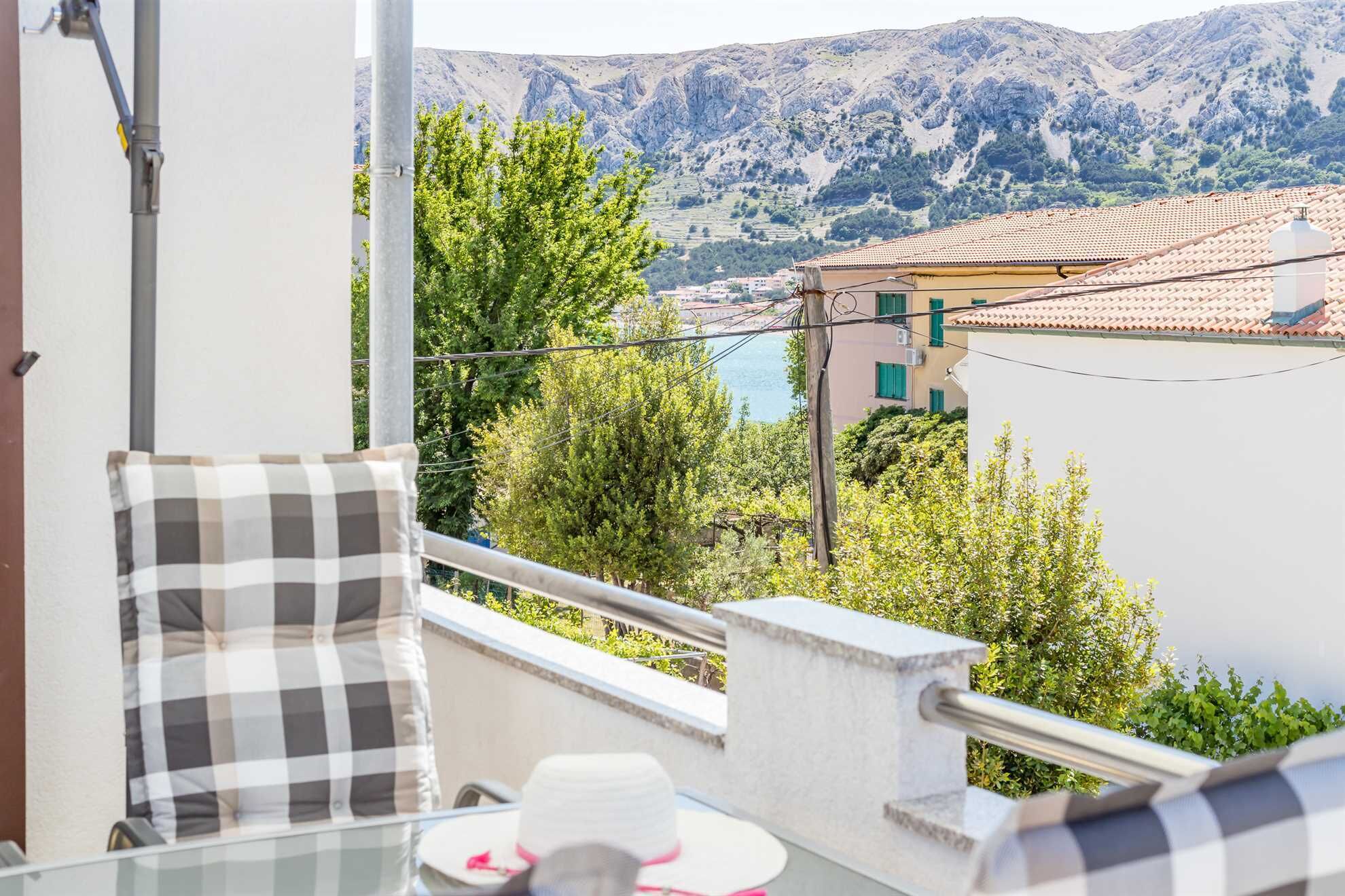 A balcony with a checkered cushion, a table with a hat, and a view of a mountainous landscape with buildings.