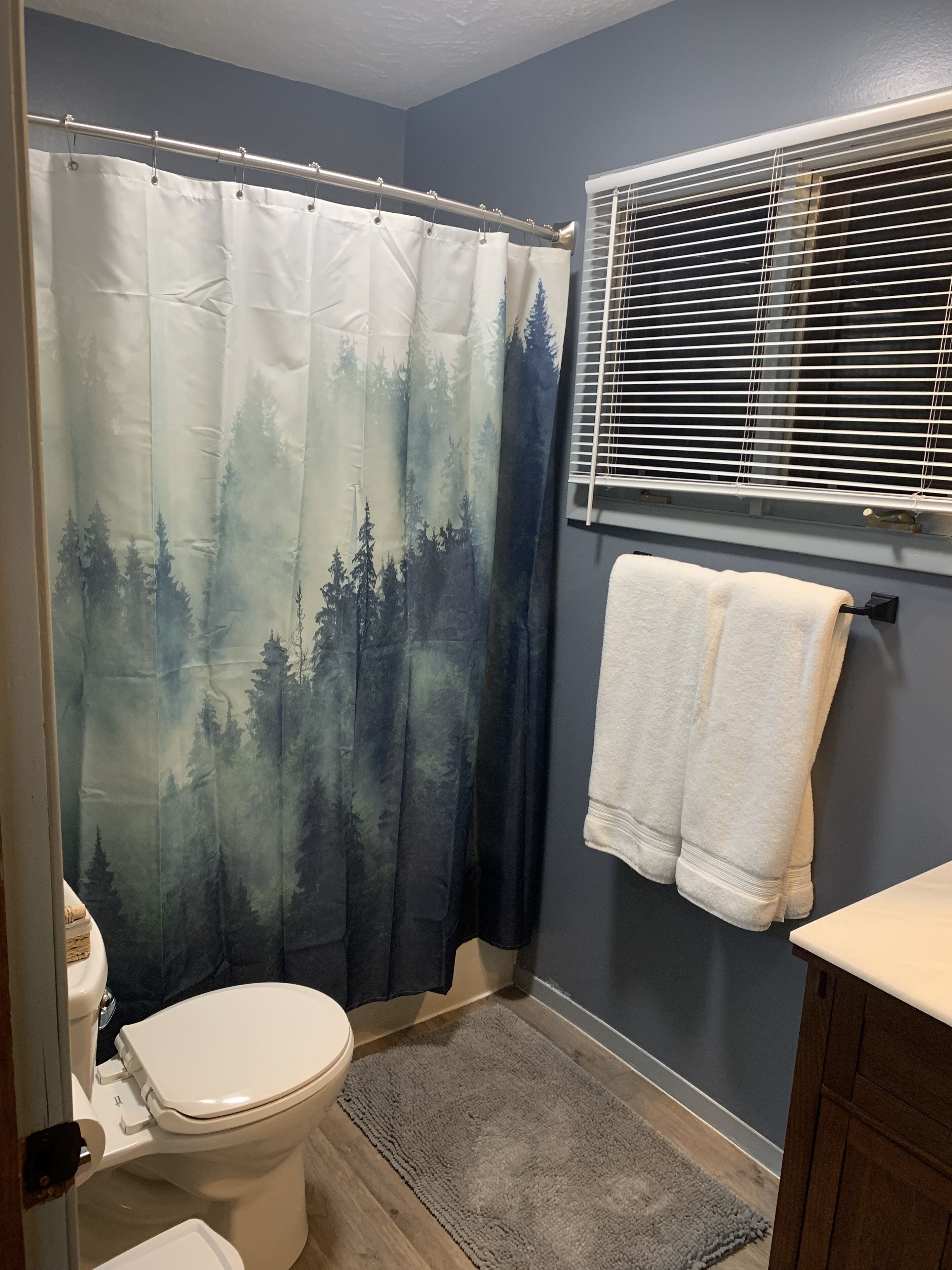 A bathroom with a shower curtain featuring a forest scene, a toilet, a sink with a towel, and a window with blinds.