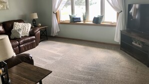 A living room with a brown leather sofa, a wooden coffee table, a TV on a wooden stand, and a window with white curtains. - Furnished Acreage by Mahoney - Sleeps 17!  Fire Pit - Shuffleboard - Clean! (Ashland)