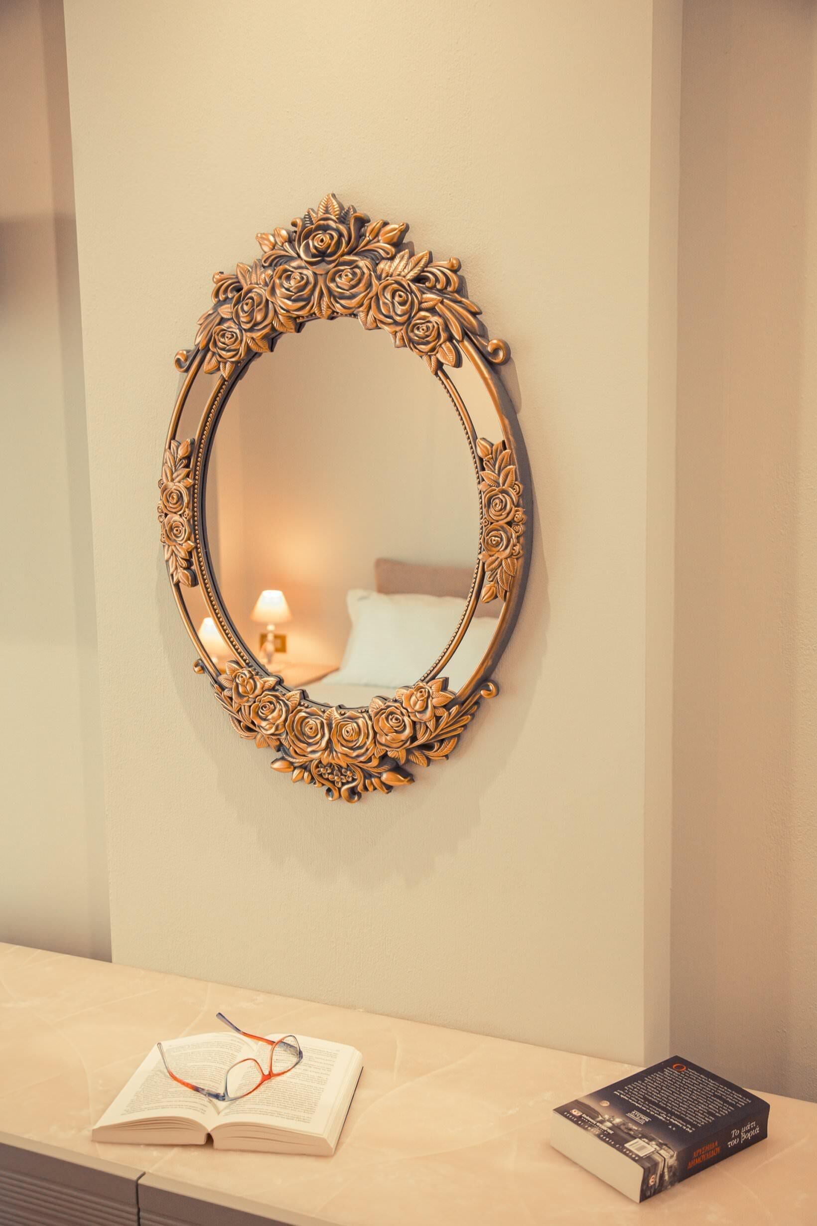 A mirror with an ornate frame on a wall, a bedside table with a book and glasses, and a lamp in the background.
