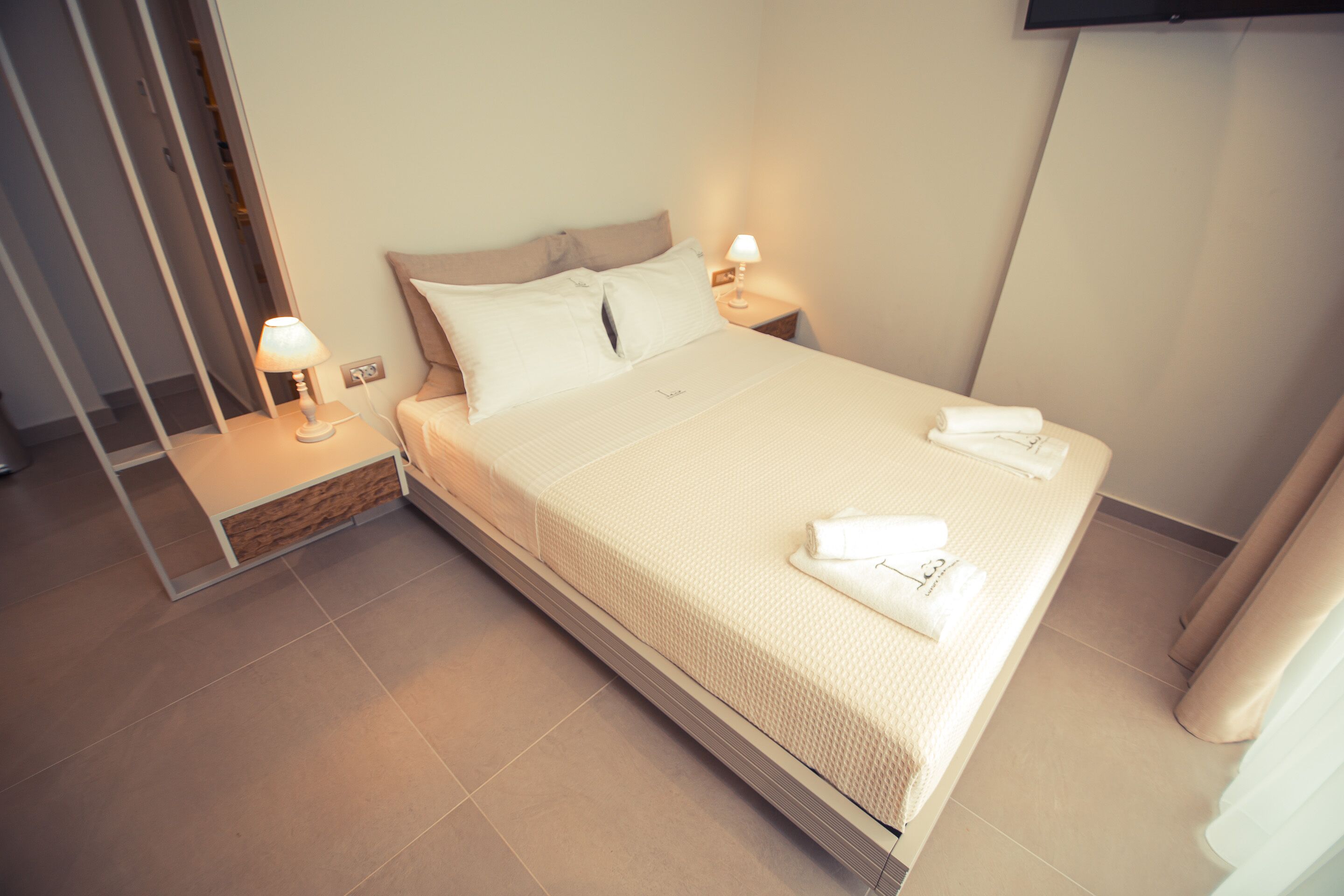 Una cama bien hecha con sábanas y almohadas blancas, una mesita de noche con una lámpara y un televisor de pantalla plana montado en la pared.