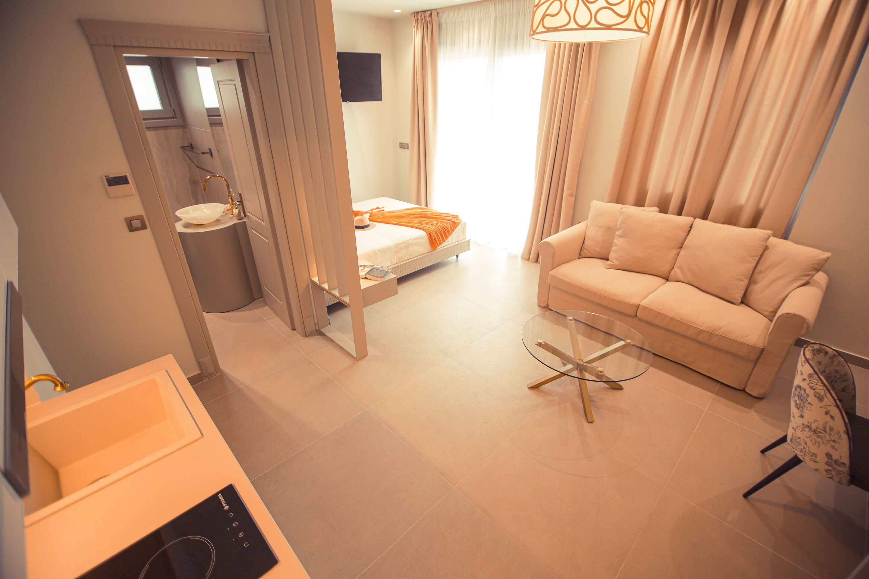 Een moderne hotelkamer met een bed, een bank, een glazen salontafel en een badkamer met wastafel en spiegel.