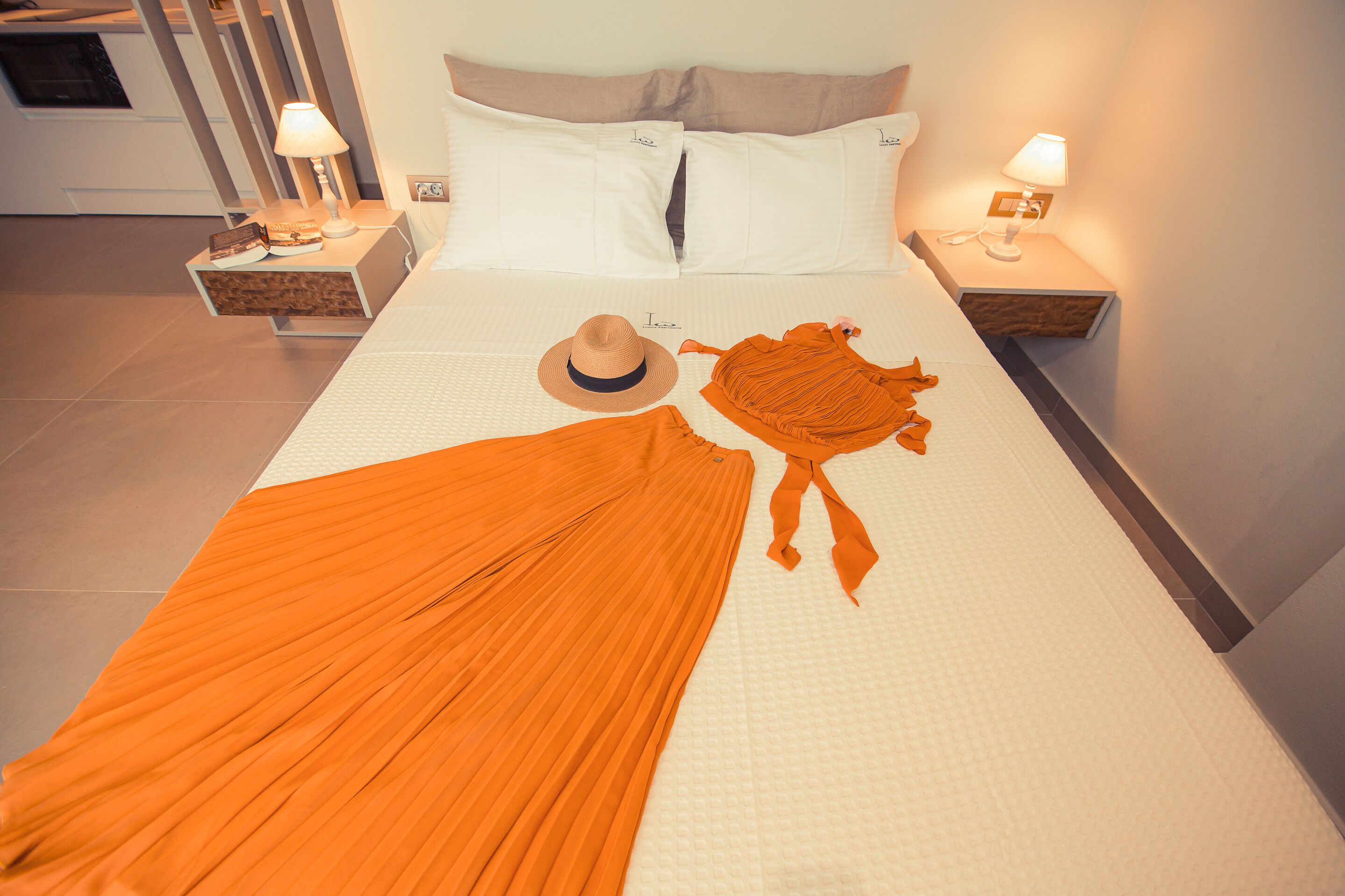A neatly made bed with a white comforter, two pillows, a hat, and an orange garment on the bed. There are two bedside tables with lamps.