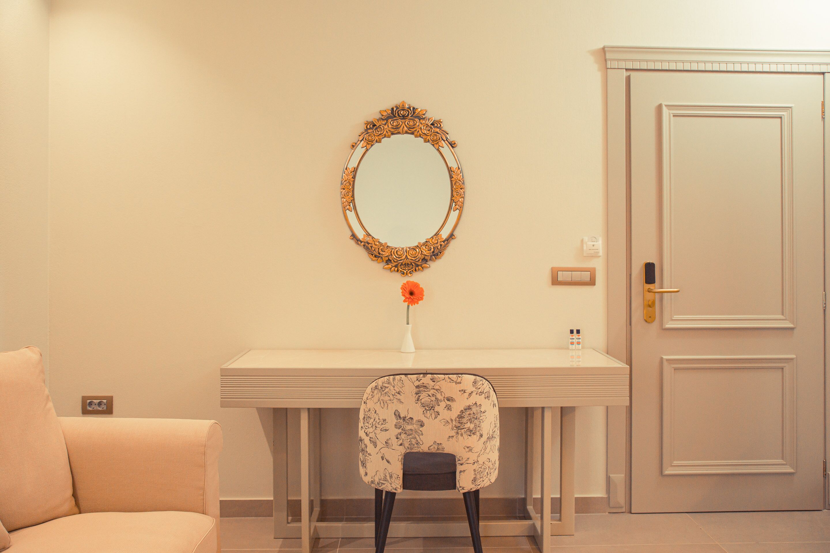 A room with a white desk, a chair with floral upholstery, a mirror with an ornate frame, and a closed door.
