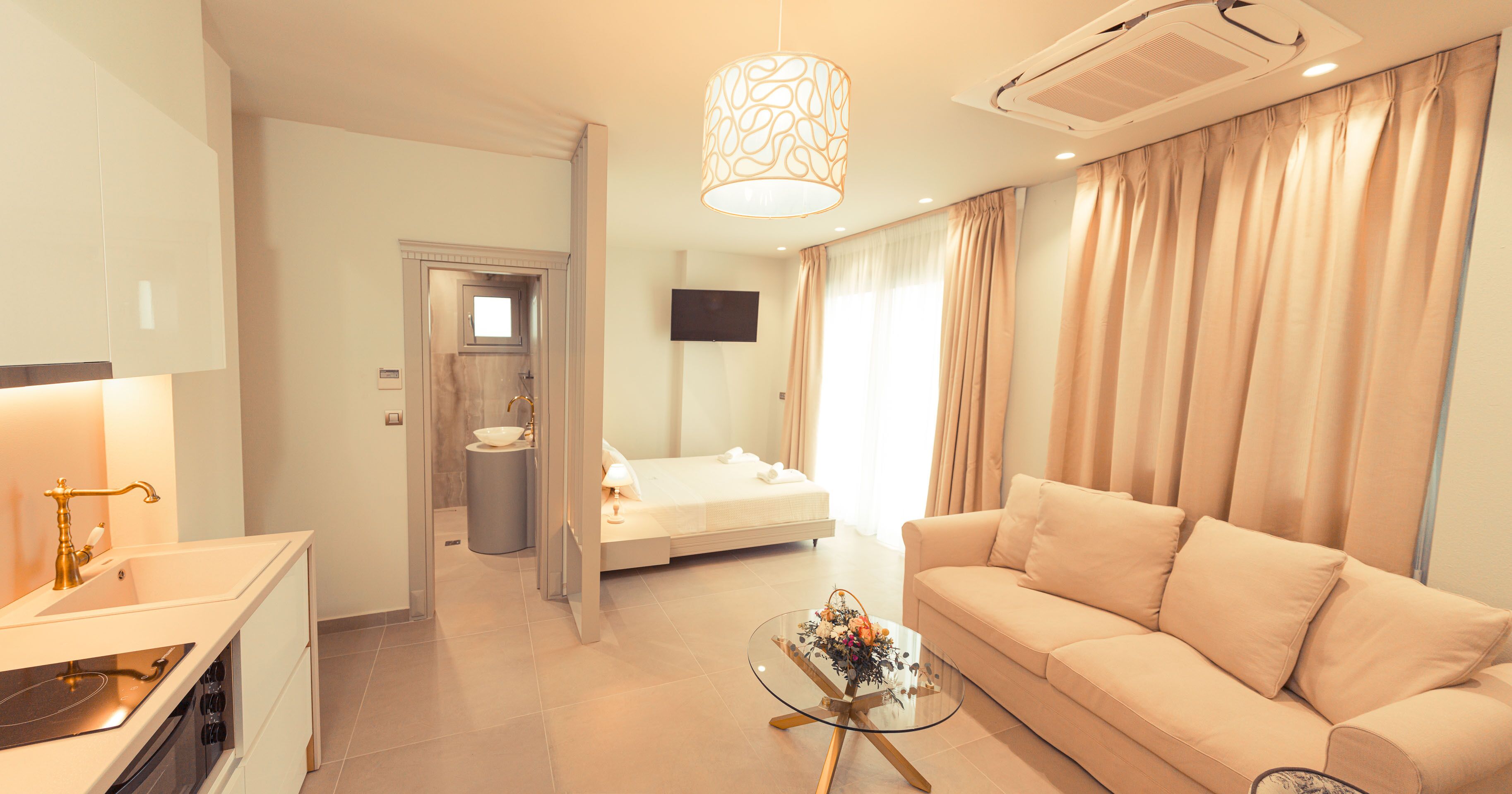 A modern hotel room with a kitchenette, a sofa, a glass coffee table, a bed, and a TV.