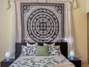 A bedroom with a large tapestry, a bed with leaf-patterned bedding, two bedside lamps, and a nightstand.