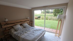 Quarto com cama de madeira, mesa de cabeceira, abajur e vista para um jardim através de uma porta de correr.