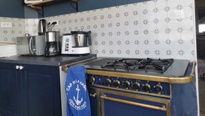 Eine Küche mit Gasherd, Kaffeemaschine und Wasserkocher auf der Arbeitsplatte. Ein blaues Handtuch hängt am Herd, darüber befindet sich ein Regal mit Gläsern und einem gerahmten Foto.