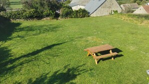 Eine grüne Wiese mit einem hölzernen Picknicktisch, Steinbauten mit Schornsteinen und einem Blick auf das Meer im Hintergrund.