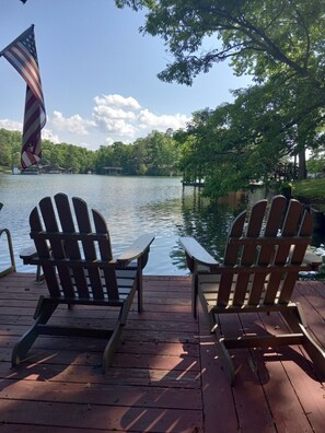 Een houten terras met Adirondack-stoelen met uitzicht op een meer, een vlag en bomen op de achtergrond.