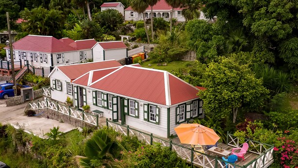 A white house with a red roof, surrounded by lush greenery and a wooden deck with outdoor furniture. - Historic Saban cottage in Windwardside, all amenities only a 2 minute walk away! (Windward Side)