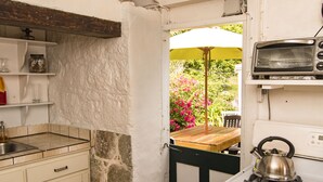 A compact kitchen with a stove, a toaster, and a view of a wooden table and pink flowers outside. - Historic Saban cottage in Windwardside, all amenities only a 2 minute walk away! (Windward Side)