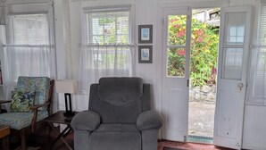 A room with a grey armchair, a wooden table, a patterned armchair, a rug, and a door leading outside. - Historic Saban cottage in Windwardside, all amenities only a 2 minute walk away! (Windward Side)