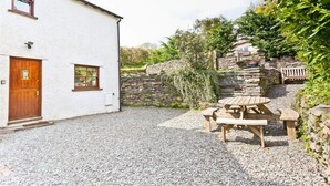 A white building with a wooden door and windows, a stone wall, and a gravel patio with a wooden table and benches. - Family-sized Lake District village house with garden, plus gorgeous walks from the door (Coniston)