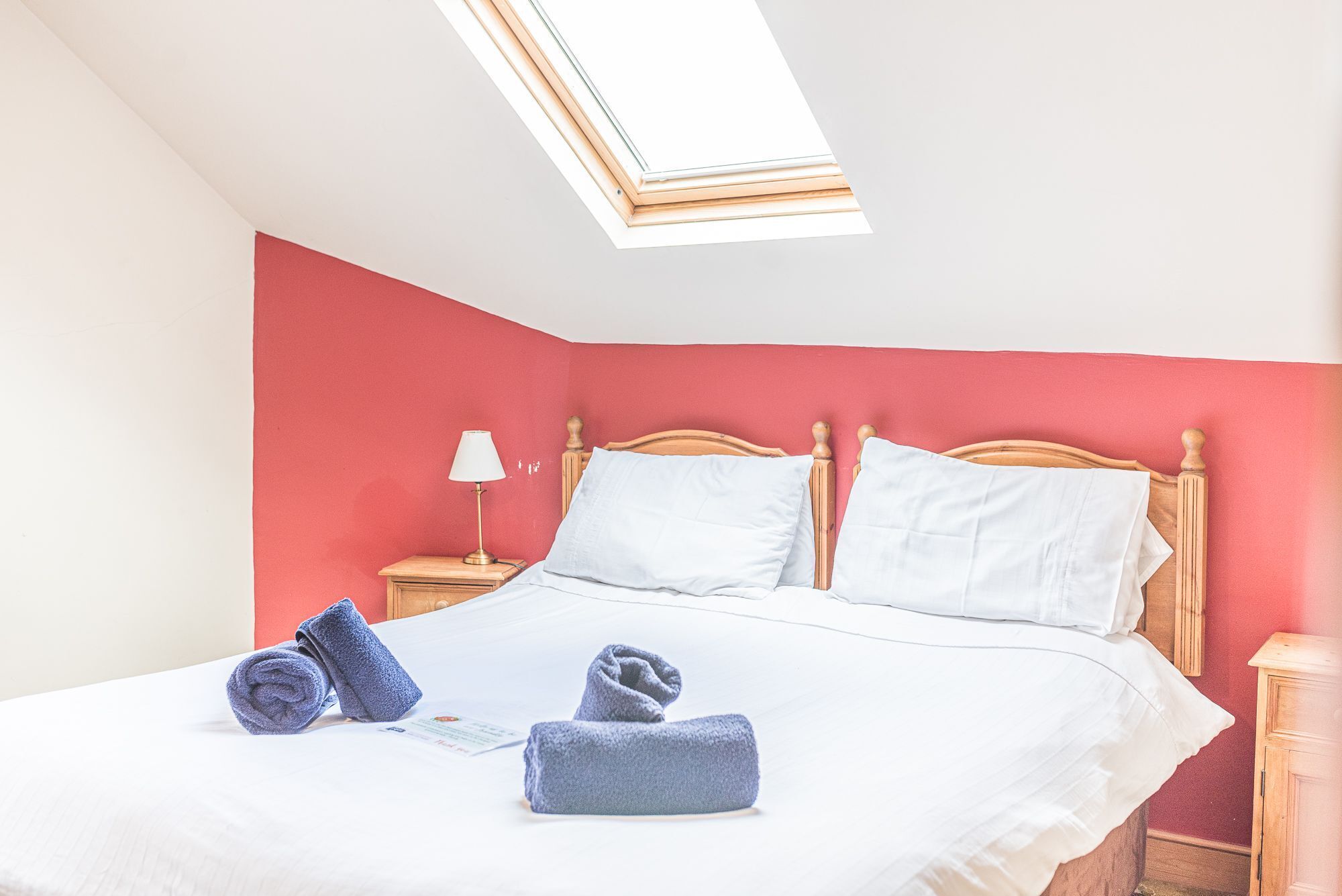 A bedroom with a bed, two pillows, a nightstand with a lamp, and two rolled towels.