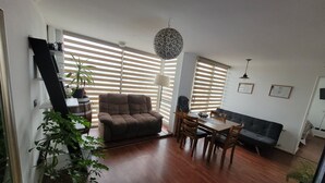 A spacious living area with a sofa, a dining table with chairs, and a large window with blinds. There is a ceiling light fixture and a potted plant. - EXELENTE VISTA AL MAR Y MUY CERCANOS A PLAYAS, CENTRO DE LA CIUDAD. (Valparaíso)