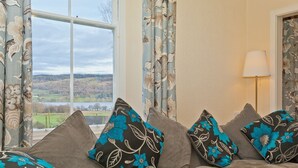 A sofa with floral cushions, a lamp, and a view of a lake and hills through a window. - Large Lake District House in popular village with gorgeous lake views (Coniston)