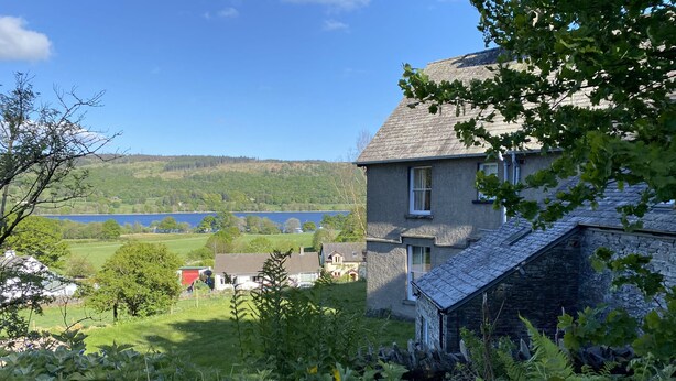 A house with a stone exterior, a sloped roof, and a view of a lake and hills in the background. - Large Lake District House in popular village with gorgeous lake views (Coniston)