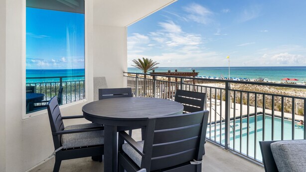 A balcony with a round table and chairs, a view of the beach, and a swimming pool.