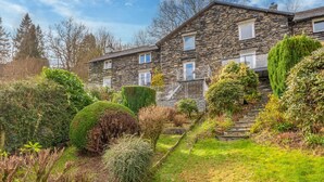 Ein Steinhaus mit zahlreichen Fenstern und einem gepflegten Garten mit verschiedenen Sträuchern und einer Rasenfläche.