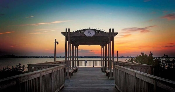 A wooden pier leading to a gazebo with a circular emblem, overlooking a body of water at sunset.