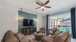 A modern living room with a ceiling fan, a flat-screen TV, and a view of a pool through large windows. - Private Pool, Spa and Premium Resort Amenities Included! (Kissimmee)