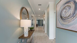 A hallway with a marble floor, a wooden console table with a white lamp, a large mirror, and a framed abstract painting. - Private Pool, Spa and Premium Resort Amenities Included! (Kissimmee)