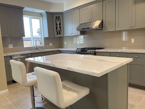 A modern kitchen with grey cabinets, a central island with bar stools, a stainless steel range hood, and a window providing natural light. - Cozy spacious house near RoseBowl, DTLA, and more (Pasadena)