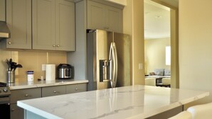 Una cocina moderna con gabinetes grises, un refrigerador de acero inoxidable, una encimera de mármol y vistas a un área de estar con un sofá y una mesa de centro.