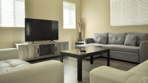 Una sala de estar moderna con televisor de pantalla plana, sofá gris, sillones blancos y una mesa de centro.