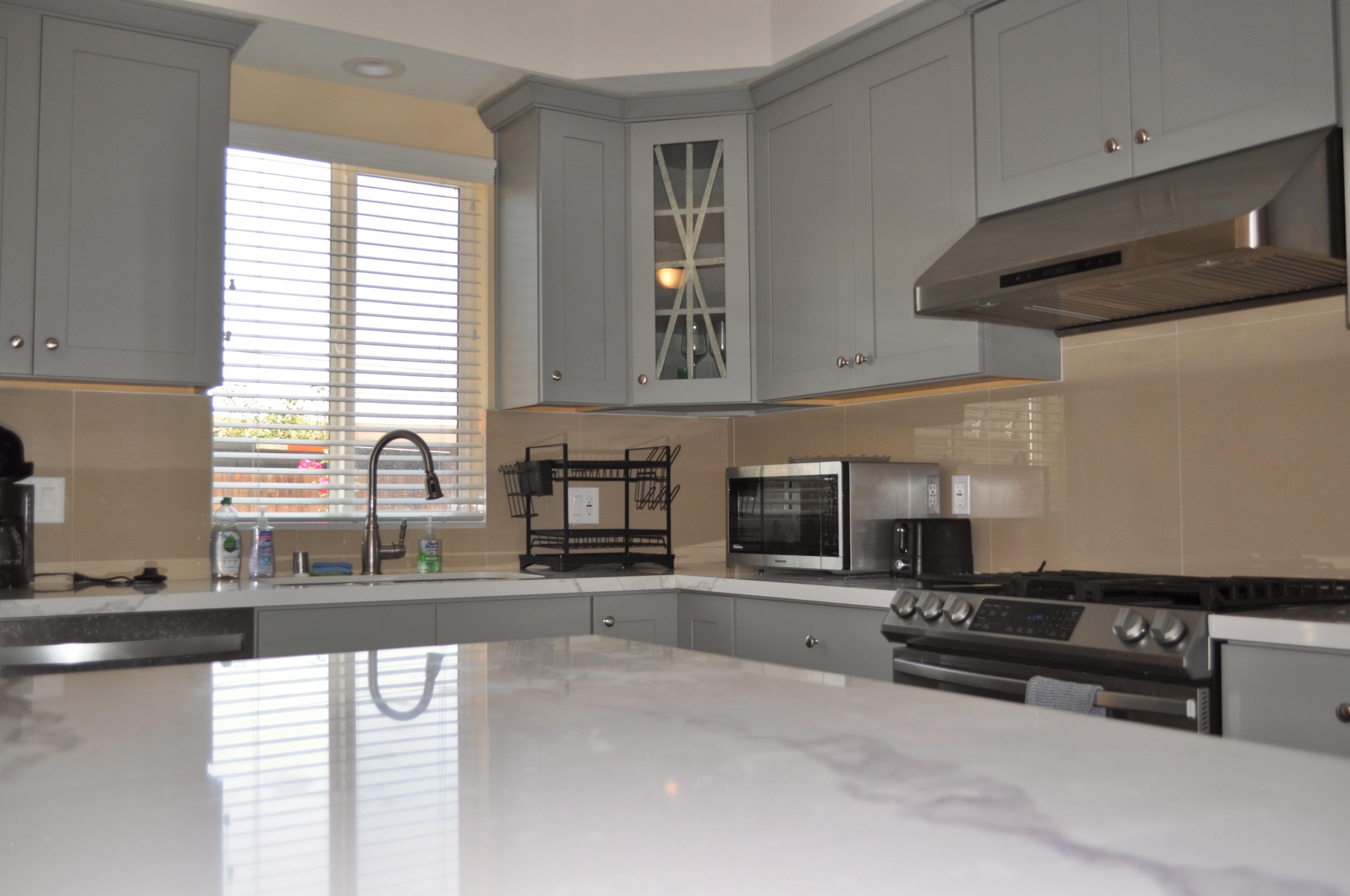 A modern kitchen with grey cabinets, a large island, stainless steel appliances, and a window with blinds.