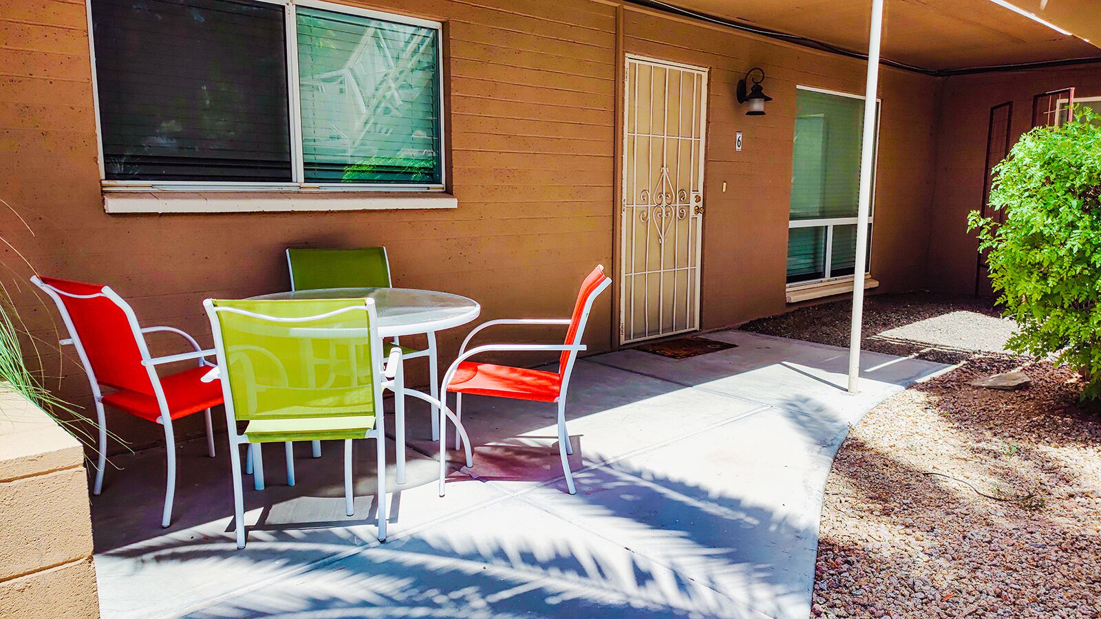 An outdoor patio with a table and four chairs, a small plant, and a building with a window and a door.