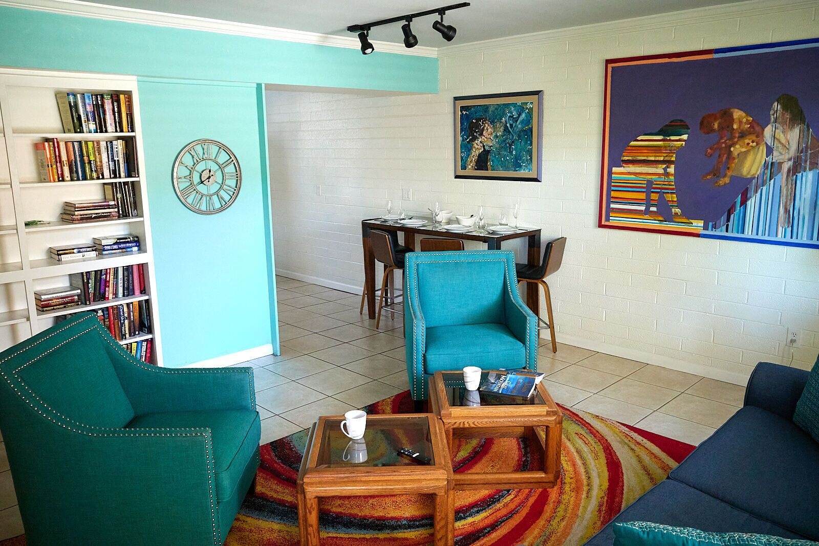A living room with teal sofas, a coffee table, a bookshelf, a dining area, and a colorful abstract painting.