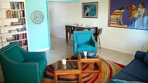 A living room with teal sofas, a coffee table, a bookshelf, a dining area, and a colorful abstract painting.