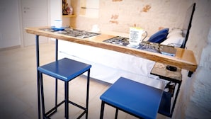 A small kitchenette with a wooden countertop, two blue stools, and a bed in the background.