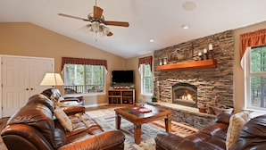 A living room with leather sofas, a stone fireplace, a ceiling fan, and a television. - 164 Jillian - Private, Peaceful Near Deschutes River, Hot Tub, Fenced Yard, Ping Pong, & Foosball (Sunriver)