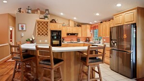 A kitchen with wooden cabinets, a stainless steel refrigerator, and a central island with bar stools. - 164 Jillian - Private, Peaceful Near Deschutes River, Hot Tub, Fenced Yard, Ping Pong, & Foosball (Sunriver)
