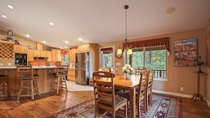 A spacious kitchen with wooden cabinets, a dining area with a wooden table and chairs, and a large window with a view of trees. - 164 Jillian - Private, Peaceful Near Deschutes River, Hot Tub, Fenced Yard, Ping Pong, & Foosball (Sunriver)
