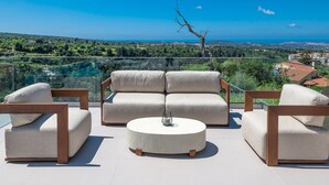 A modern outdoor seating area with a sofa and chairs, a round table, and a view of a coastal landscape. - Dim Luxury Villa - With Private Pool (Margarites)