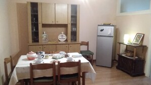 Un comedor con una mesa preparada para cuatro, sillas de madera, un armario con puertas de vidrio, un refrigerador y un pequeño estante con objetos decorativos.
